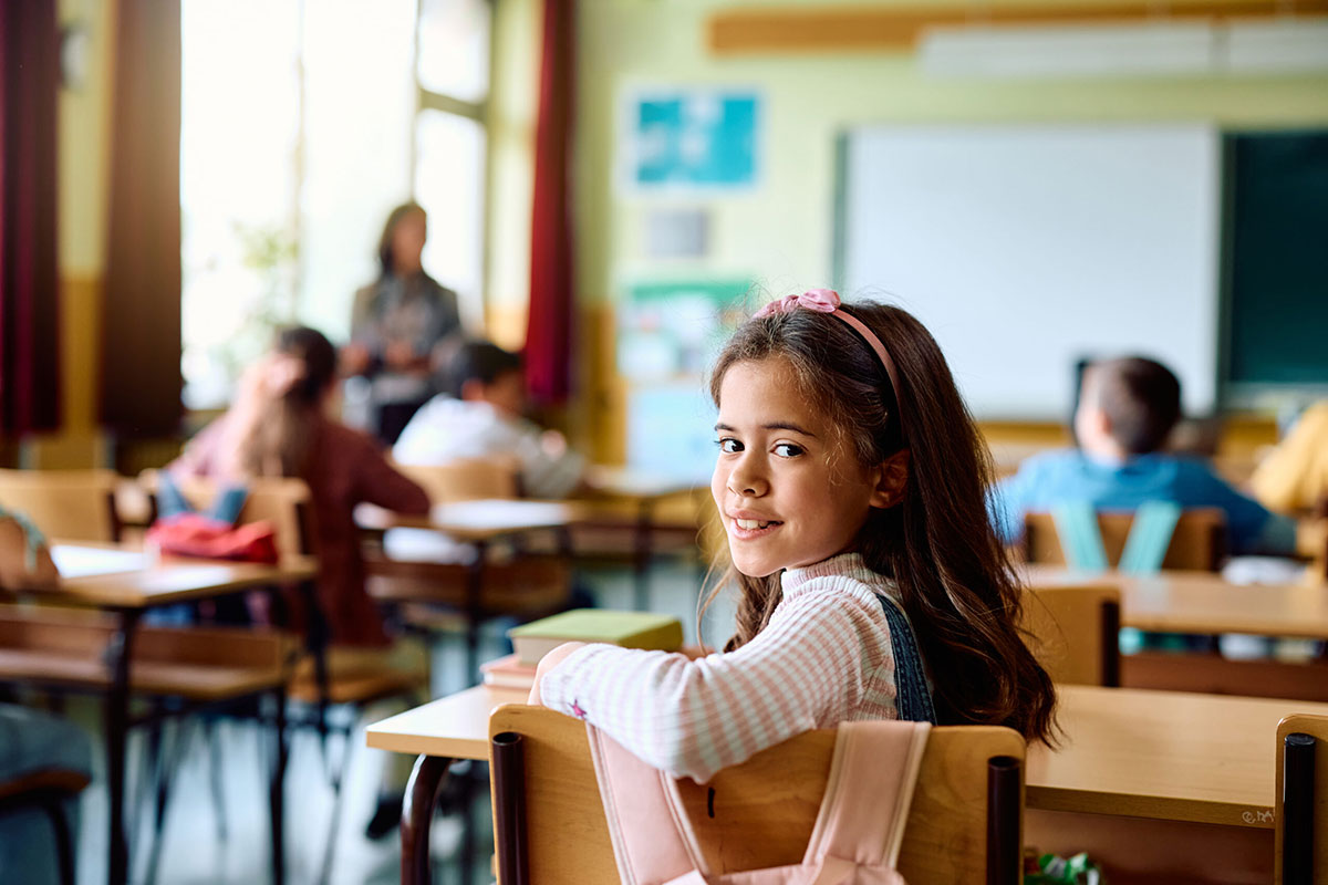 A picture of a young girl smiling in the classroom sitting at a desk for a blog about the link between kids smiles and their academic performance